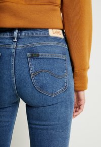 Blue denim jeans with a fitted design, visible stitching, and two back pockets. Lee logo tag on waistband, textured fabric, medium rise.