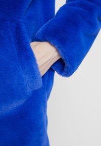 Faux fur blå jacka med en mjuk textur där en hand placeras i en sidoficka, som visar plaggets livliga färg och design.