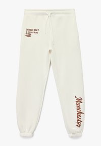 Pantalones de chándal blancos con cinturilla ajustable. Presenta texto impreso en marrón, incluyendo "Manchester" y un diseño de fecha/dirección. Puños elásticos.