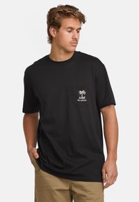 Camiseta de algodón negra con mangas cortas, con un bolsillo en el pecho y un diseño de palmera blanca. La tela es suave y transpirable.