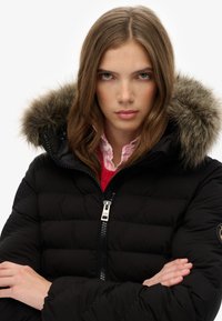 Chaqueta acolchada negra con capucha rematada en piel, cierre de cremallera y textura acolchada. Se lleva sobre un suéter rojo con una camisa rosa de cuello debajo.