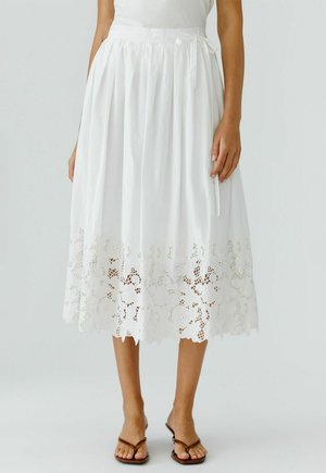 Weißer Midirock mit Spitzenabschluss und gebundener Taille, getragen mit braunen Sandalen mit offenen Zehen.