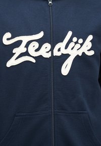 Laivastonsininen vetoketjullinen collegepaita, jossa valkoiset kursiivikirjaimet "Zeeldijk" rinnassa ja etutaskut näkyvissä alareunassa.