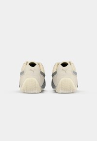 Beige Puma sneakers med teksturerede gummisåler og grå stribedetaljer, set fra bagsiden på en ensfarvet hvid baggrund.