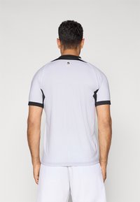 Mann mit kurzen dunklen Haaren, der ein weißes Sporttrikot mit schwarzem Rand trägt und mit dem Rücken zu einer einfachen hellen Hintergrund steht.