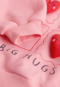 Pink sweatshirt med ribbede manchetter, prydet med broderede røde hjerter og teksten "Big HUGS" i fede sorte bogstaver på blødt stof.