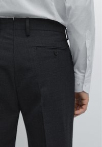 Dunkelgraue, maßgeschneiderte Hose aus strukturiertem Stoff, mit einer einzelnen Gesäßtasche mit Knopfverschluss, kombiniert mit einem weiß gestreiften Hemd.