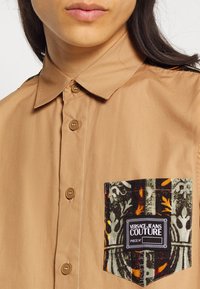 Ljusbrun skjorta med krage och knappar, med en dekorativ mönstrad ficka med flerfärgade detaljer och en etikett.