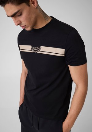 Mężczyzna z tatuażem na szyi patrzy w dół, ubrany w czarny t-shirt z beżowymi paskami na klatce piersiowej i logo "Sport Club Guess", ręce w kieszeniach.