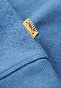 Tejido de sudadera azul con una textura suave, que presenta una etiqueta amarilla cosida en el lado. Detalle de costura visible a lo largo del borde.