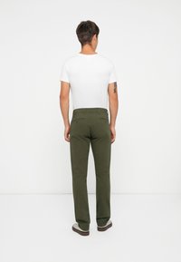 Green chinos made of cotton, featuring a straight leg design, back pockets with zippers, worn with a white T-shirt and sneakers.