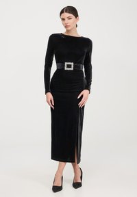 Abito in velluto nero con maniche lunghe, silhouette aderente, spacco fino alla coscia e una cintura larga con una grande fibbia di strass.