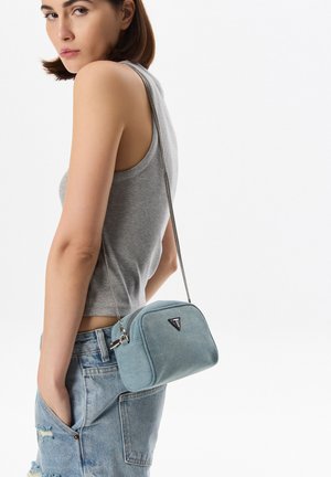 Modrá denimová crossbody taška se stříbrným řetízkovým popruhem, obdélníkového tvaru, s logem a texturovaným povrchem, nošená přes šedé tílko.