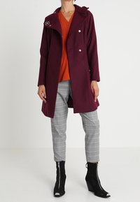 Burgundy kabát s kapucí a designem na dvojité zapínání. Šedé kostkované kalhoty a černé kotníkové boty se zipy. Jasně oranžová blůza pod ním.