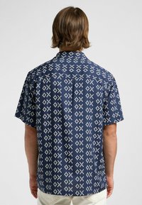 Chemise à manches courtes, couleur marine, ornée d'un motif géométrique blanc. Le tissu est doux, avec un col et un ourlet droit.