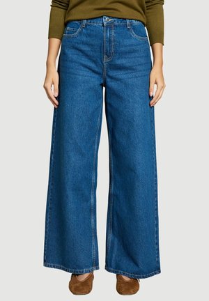 Person trägt hoch taillierte, weit geschnittene blaue Jeans kombiniert mit braunen flachen Schuhen und einem olivgrünen Langarmshirt.