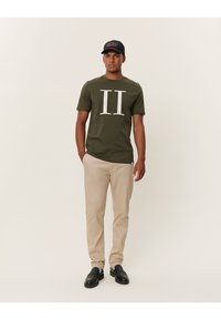 Oliwkowa bawełniana koszulka z białym nadrukiem "II", zestawiona z beżowymi chinosami i czarnymi butami. Czapka jest czarna z różowym napisem.