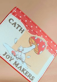 Cartera roja con acentos de lunares, superficie texturizada y una ilustración de un oso con un hongo y flores. El texto dice "CATH JOY MAKERS."