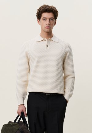 Jeune homme aux cheveux bouclés portant une chemise crème texturée à manches longues et un pantalon noir, tenant une casquette verte et un sac sombre à ses côtés.
