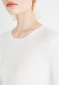 Un pull en tricot blanc avec un col rond, présentant un motif texturé. Le tissu est léger et légèrement transparent, avec des manches dolman.