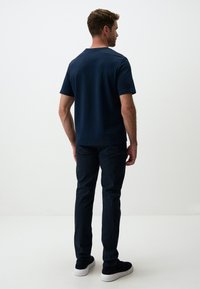 T-shirt navy a maniche corte con scollatura rotonda, abbinato a pantaloni scuri e scarpe scure, visto da dietro su uno sfondo chiaro.