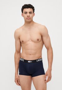 Sötétkék férfi boxeralsó, puha anyagból készült, kontrasztos fekete derékkal, amelyen fehér "LACOSTE" felirat és logó található.