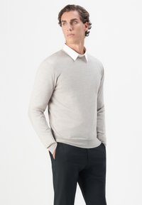 Nuori mies, jolla on kiharat hiukset, pukeutuneena vaaleanharmaaseen villapaitaan, valkoisen kauluspaidan päälle, ja mustiin housuihin. Hän seisoo kädet taskuissa ja katsoo vasemmalle.