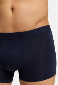 Gros plan sur le bas du torse d'un homme portant un boxer Hanro bleu marine avec une ceinture élastique visible sur un fond uni.