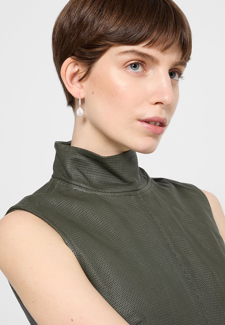 Top senza maniche verde metallico con colletto alto, caratterizzato da una superficie texturizzata e cuciture visibili, abbinato a un orecchino argento a forma di goccia.