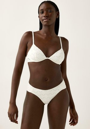 Tummaihoinen nainen valkoisessa, teksturoidussa bikinitopissa ja -housuissa, seisoo vaaleaa yksiväristä taustaa vasten, katsoo suoraan eteenpäin rentoutuneessa asennossa.