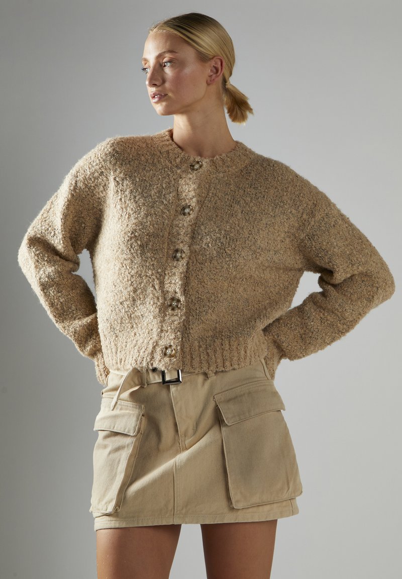 Beige gestrickte Strickjacke mit strukturiertem Finish, großen Knöpfen und geripptem Saum, kombiniert mit einem khakifarbenen Cargo-Rock mit vorderen Taschen.