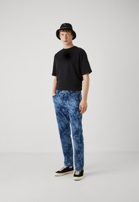 Camiseta negra de manga corta combinada con pantalones vaqueros azul jaspeado. El modelo lleva un sombrero negro de bucket y zapatos negros con cordones y suela blanca.