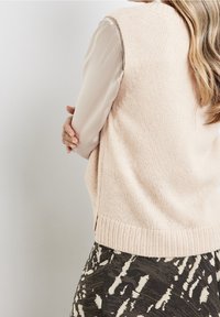 Donna con capelli lunghi che indossa un gilet di maglia beige su una blusa chiara e una gonna a righe in bianco e nero, in piedi con le braccia incrociate.