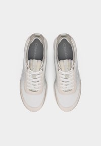 Ζευγάρι λευκά sneakers Calvin Klein με ανοιχτό μπεζ λεπτομέρειες, λευκά κορδόνια και γκρι πάτους με το λογότυπο, φωτογραφημένα από πάνω σε μονόχρωμο φόντο.