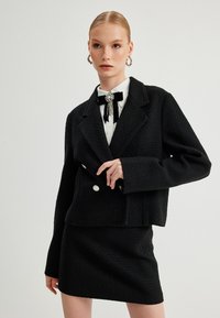 Schwarzes strukturiertes Blazer mit cropped Schnitt, doppelt geknöpften Vorderseite, passendem Rock und einer weißen Bluse, die durch eine schwarze Fliege-Brosche akzentuiert ist.