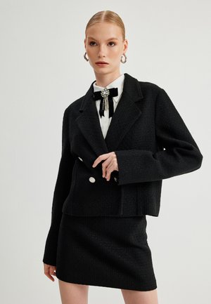 Zwarte gestructureerde blazer met een cropped fit, dubbele knopen aan de voorkant, bijpassende rok en een witte blouse voorzien van een zwarte strikspeld.