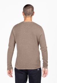 Braunfarbener Longsleeve mit gerippter Textur, rundem Ausschnitt und eng anliegenden Ärmeln. Wird mit dunklen Hosen getragen, auf denen ein seitliches Markenlabel zu sehen ist.