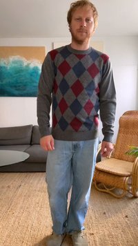 Suéter cinza em padrão argyle com diamantes em marrom e azul, usado sobre jeans azul claro. O cenário inclui uma cadeira de vime e um sofá.