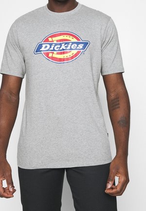 Mies, joka on pukeutunut harmaaseen Dickies-logopaitaan ja mustiin housuihin, esittelee tatuointejaan kyynärvarressaan valkoista taustaa vasten.