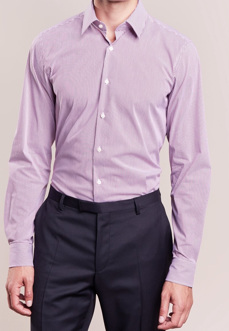 Ljuslila randig skjorta med lång ärm och spetsig krage, i kombination med mörka skräddarsydda byxor. Har vita knappar.