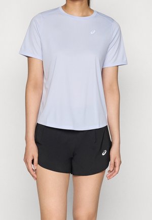 Femme portant un t-shirt gris clair à manches courtes et un short de sport noir avec des logos ASICS sur fond blanc.