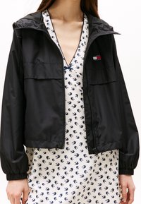 Czarna kurtka na zamek z kapturem, z dwoma przednimi kieszeniami i elastycznymi mankietami. Nosi się ją na białą sukienkę z granatowymi wzorami ptaków.