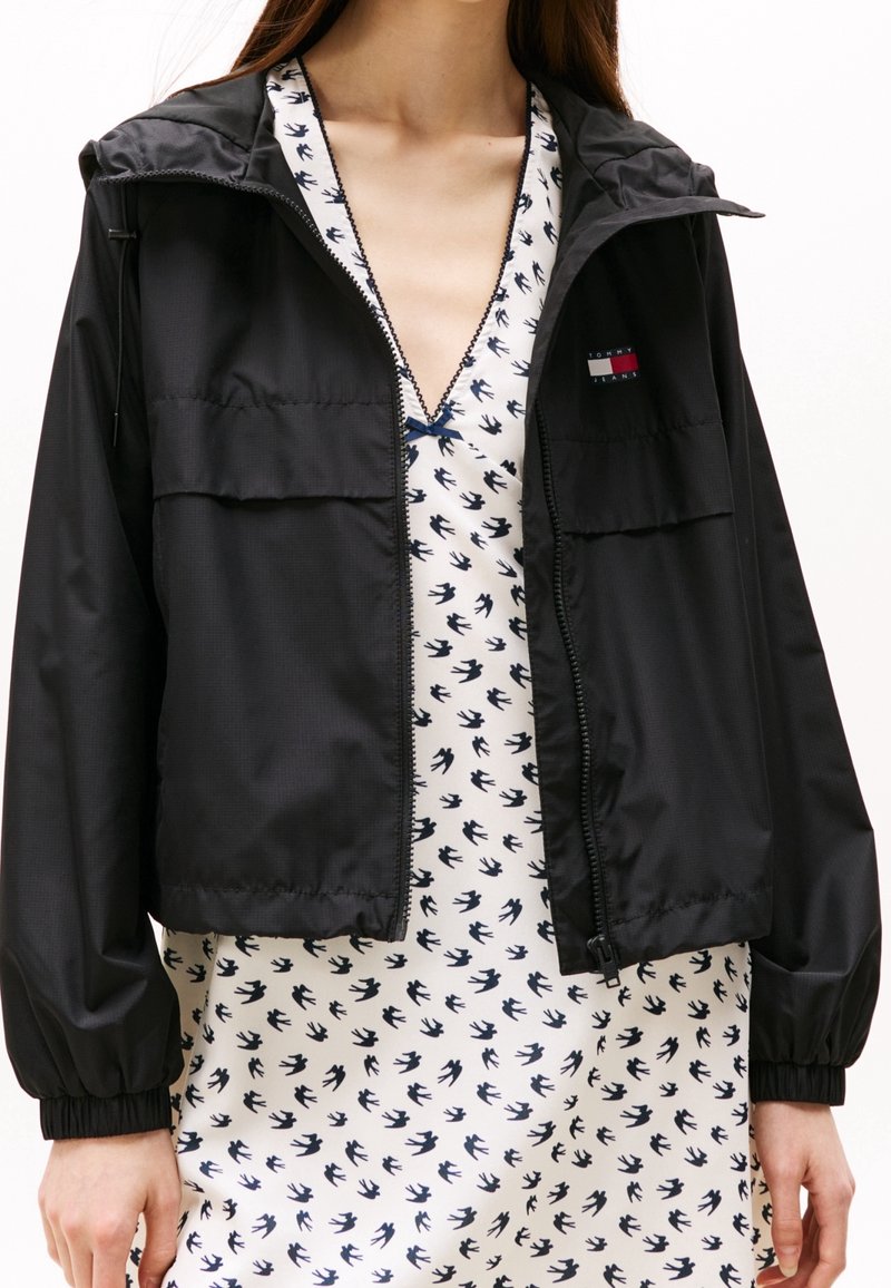 Czarna kurtka na zamek z kapturem, z dwoma przednimi kieszeniami i elastycznymi mankietami. Nosi się ją na białą sukienkę z granatowymi wzorami ptaków.