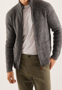 Man i en ofodrad grå stickad kofta över en vit t-shirt med olivgröna byxor, står med en hand i fickan.