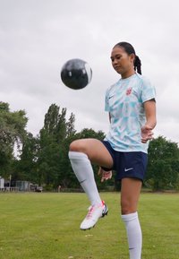 Fotbollsspelare klädd i en ljusblå, mönstrad tröja med diamantmönster, marinblå shorts, vita strumpor och svarta samt vita fotbollsskor, som jonglerar med en fotboll.