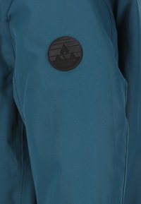 Veste de plein air teal avec une texture lisse, dotée d'un patch noir circulaire avec un design de montagne et le texte en relief "Whistler" sur la manche.