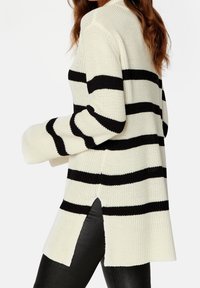Witte en zwarte gestreepte gebreide trui met lange mouwen, zijsplitsen en geribde manchetten. Materiaal lijkt textuur te hebben en zacht aan te voelen.