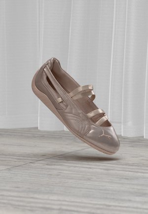 Ballerine plate métallisée or rose avec triple brides élastiques et semelle en caoutchouc, présentée sur un sol en bois clair et un rideau blanc transparent.