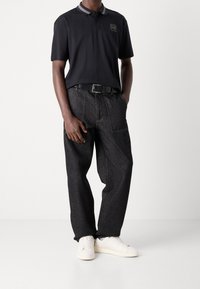 Zwarte polo met een logo, grijs gestreepte kraag, gecombineerd met donkere denim broek en witte sneakers, met zichtbare stiksels en zakken.