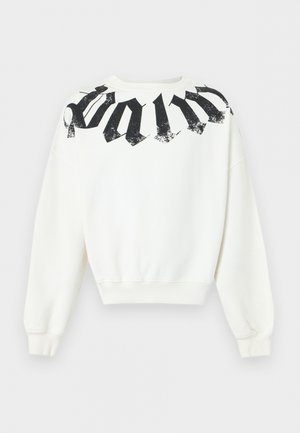 Ung mand med krøllet hår iført en hvid sweatshirt med teksten "palm", brede beige jeans og sort-hvide sneakers, står mod en hvid baggrund.